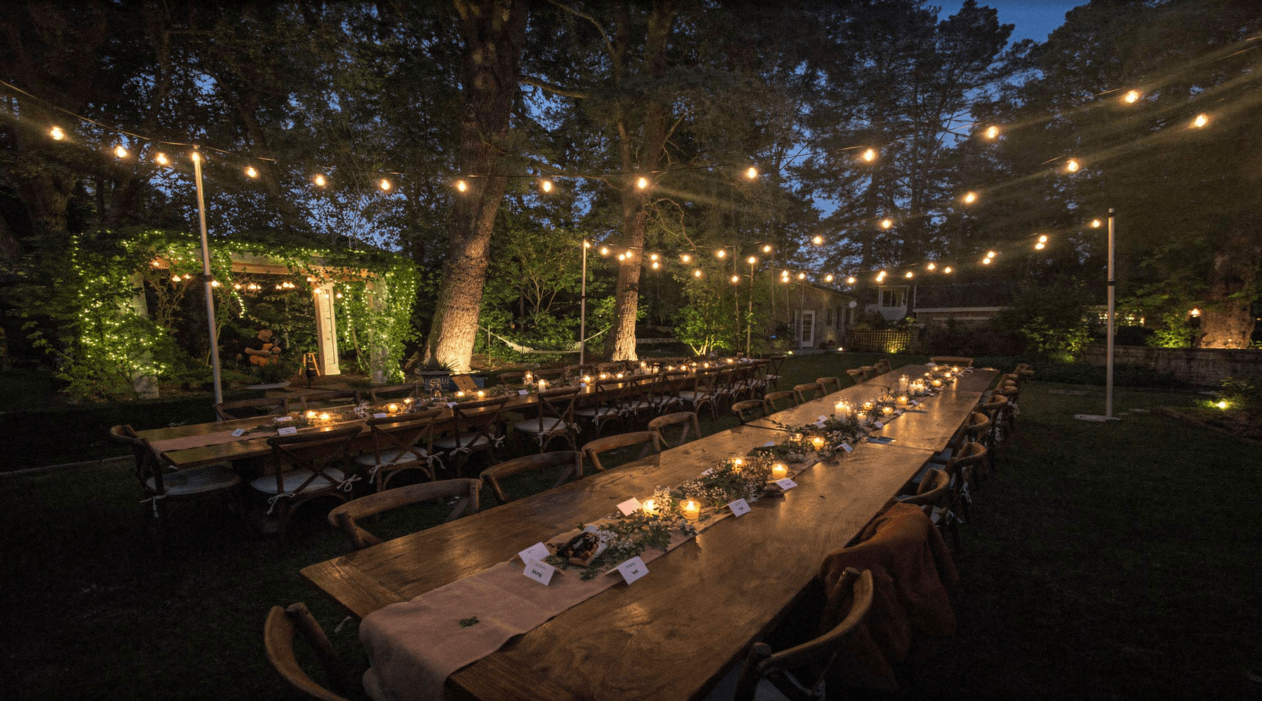 Long wooden farm tables set with candles and greenery beneath a dense warm string light canopy at a private estate dinner at dusk