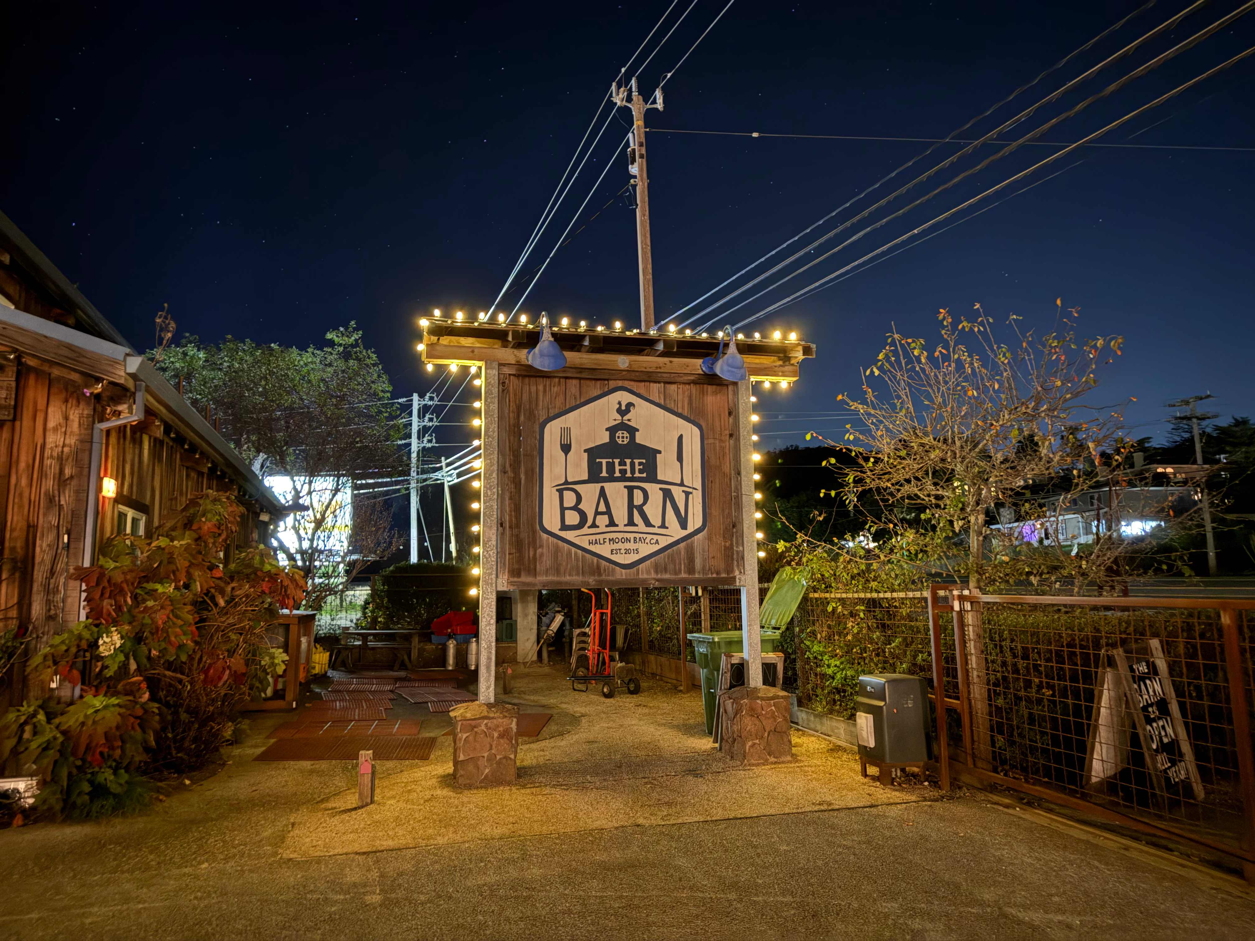The Barn restaurant entrance sign in Half Moon Bay at night, framed with warm Edison string lights against a deep blue sky