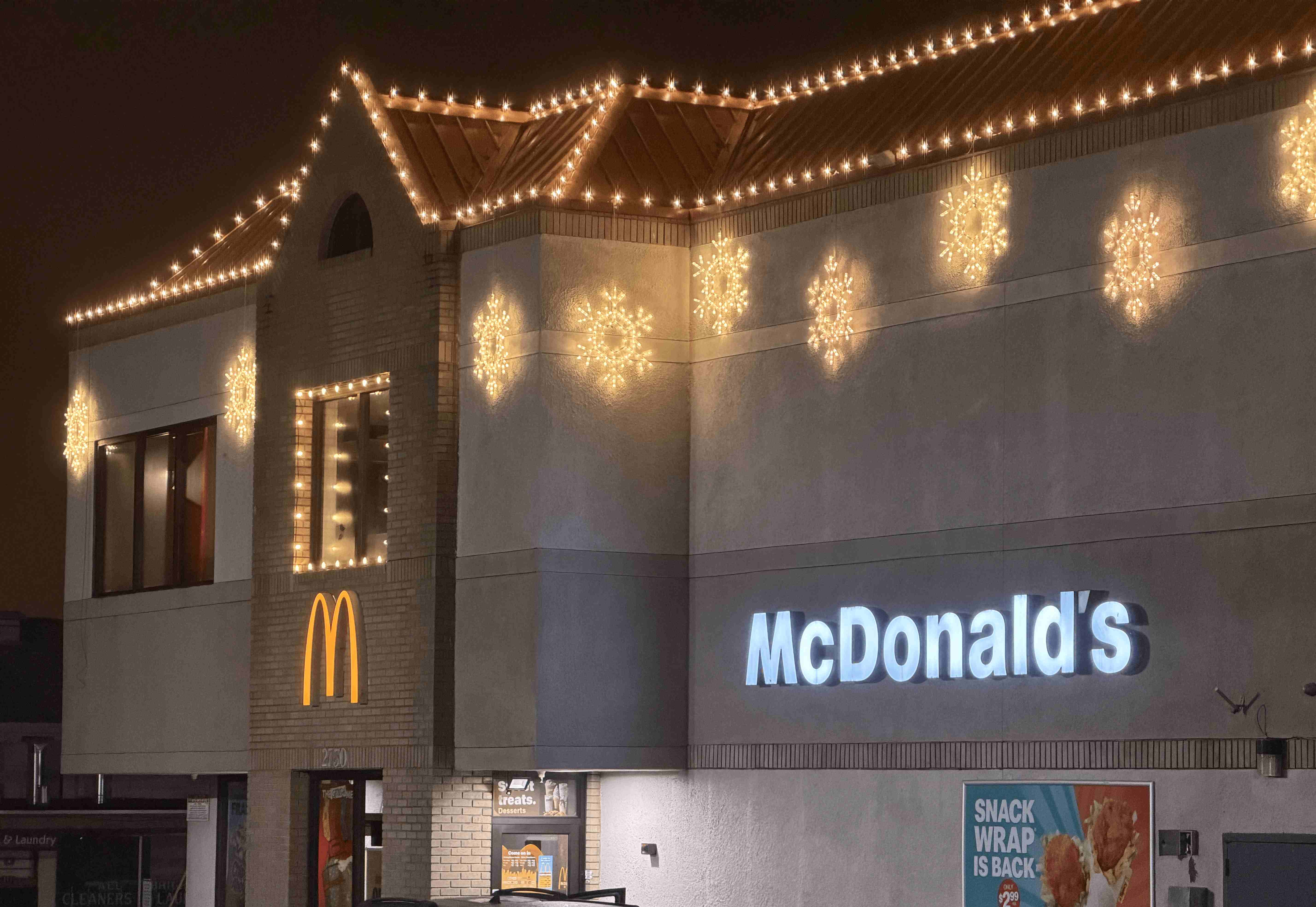 McDonald's restaurant exterior at night with warm white roofline Christmas lights and decorative snowflake medallions installed across the facade